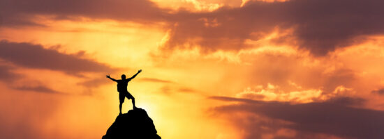 Landscape with silhouette of a standing happy man with backpack and raised-up arms on the mountain peak on the background of cloudy sky at colorful sunset in summer. Travel, Climbing, Trekking.